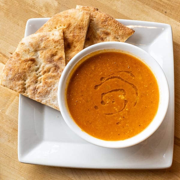 A bowl of orange soup with olive oil drizzle, served with flatbread on a white plate.