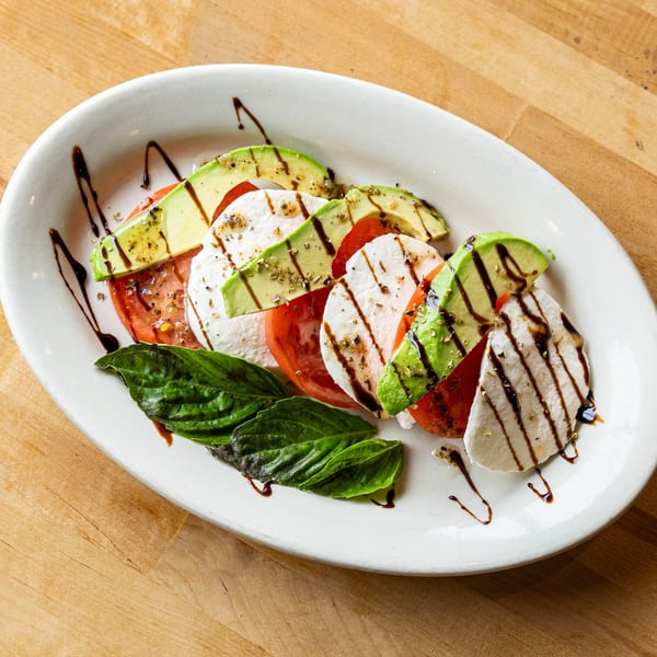Caprese salad with sliced tomatoes, mozzarella, avocado, basil, and balsamic glaze on a white plate.